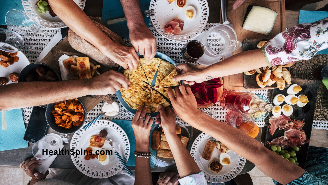 Top view of a diverse group of hands reaching for a variety of healthy foods on a shared table, highlighting the importance of eating for cancer prevention. The spread includes fruits, vegetables, whole grains, and lean proteins. HealthSpins.com